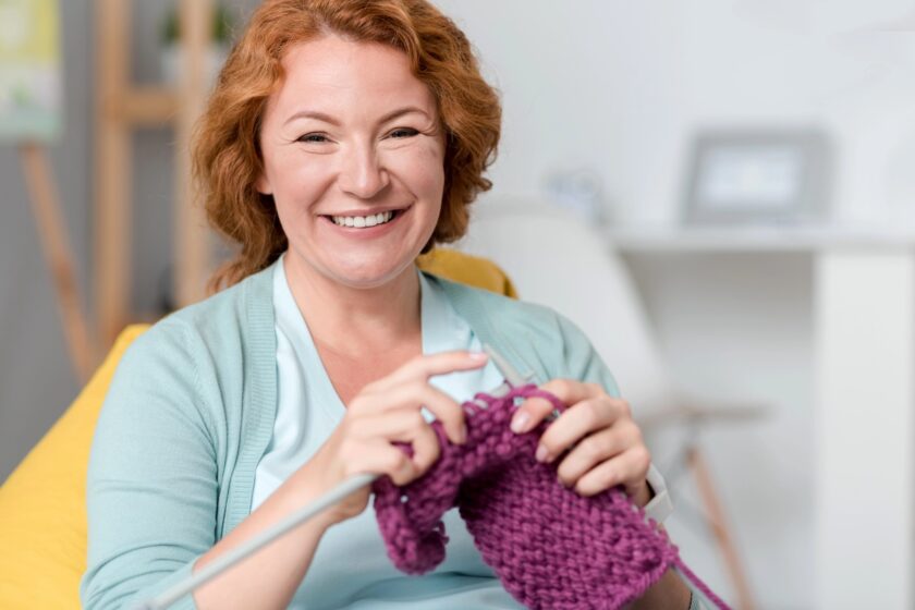 Donna seduta in poltrona mentre usa due ferri per lavorare a maglia un piccolo progetto color crema